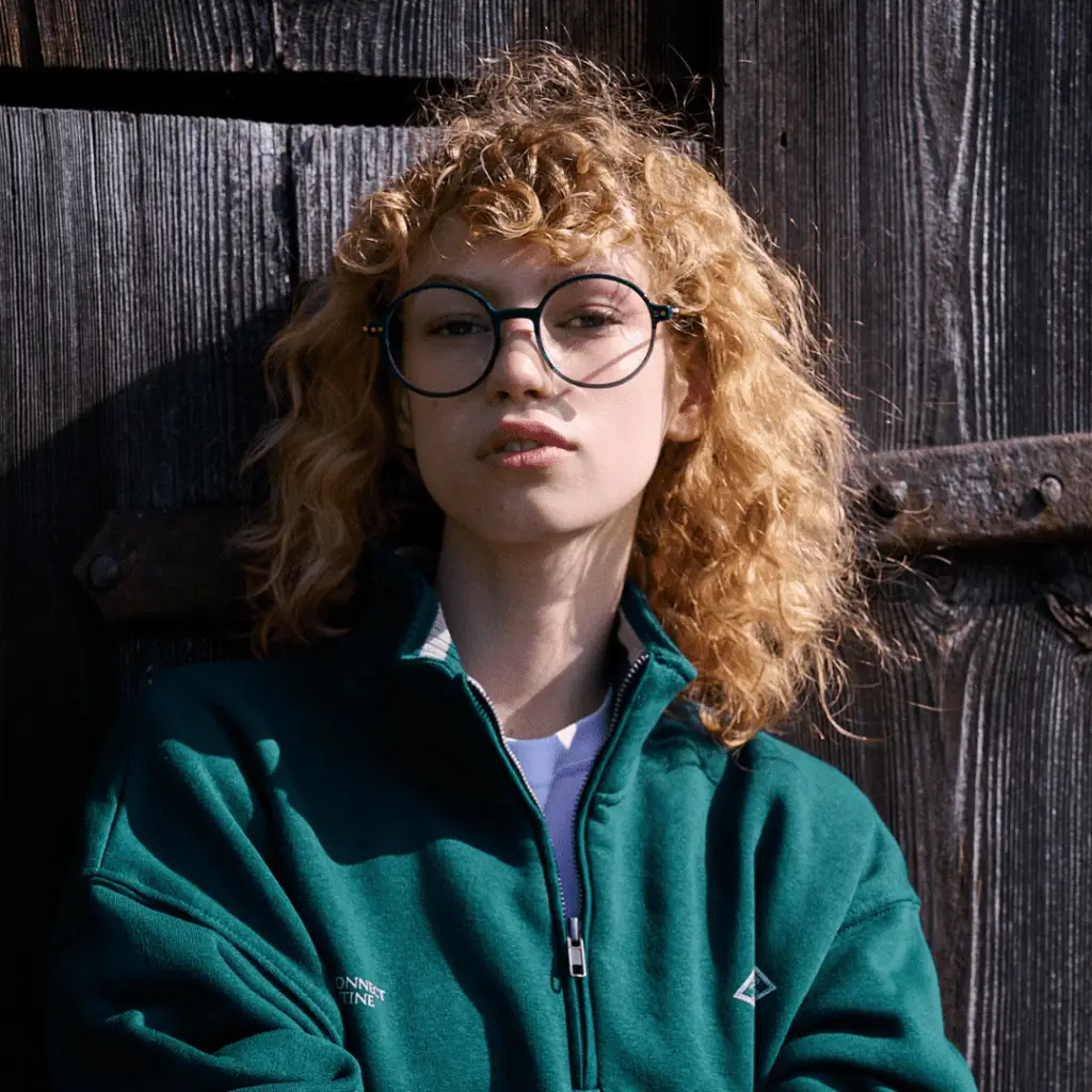 Frau mit Locken und Brille von Coblens vor Holzwand.